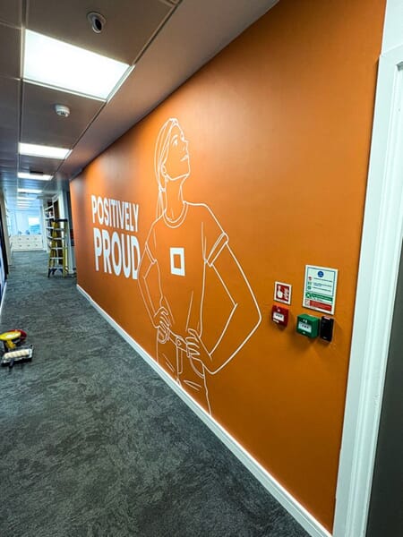 An image of the "Positively Proud" orange wall graphic, which shows a sports player looking up with a hand on their hip.